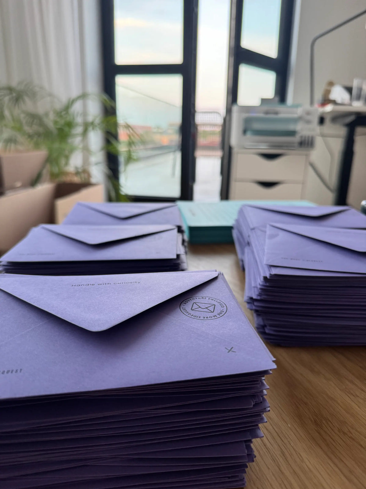 Stacks of lavender purple envelopes arranged on a wooden table in an office setting, with a window, printer, and plant visible in the blurred background.