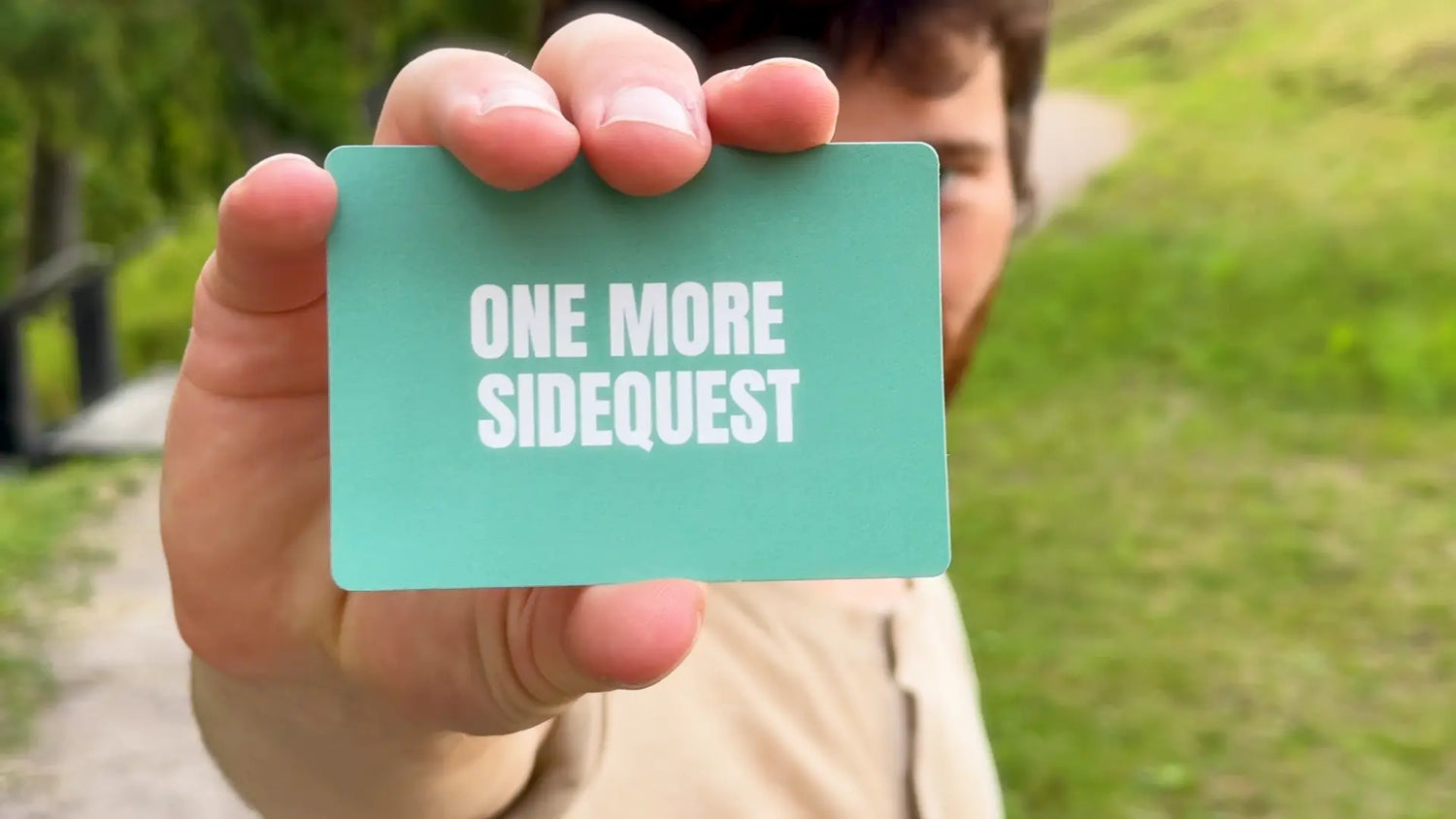 Person holding a teal card with 'ONE MORE SIDEQUEST' text outdoors.