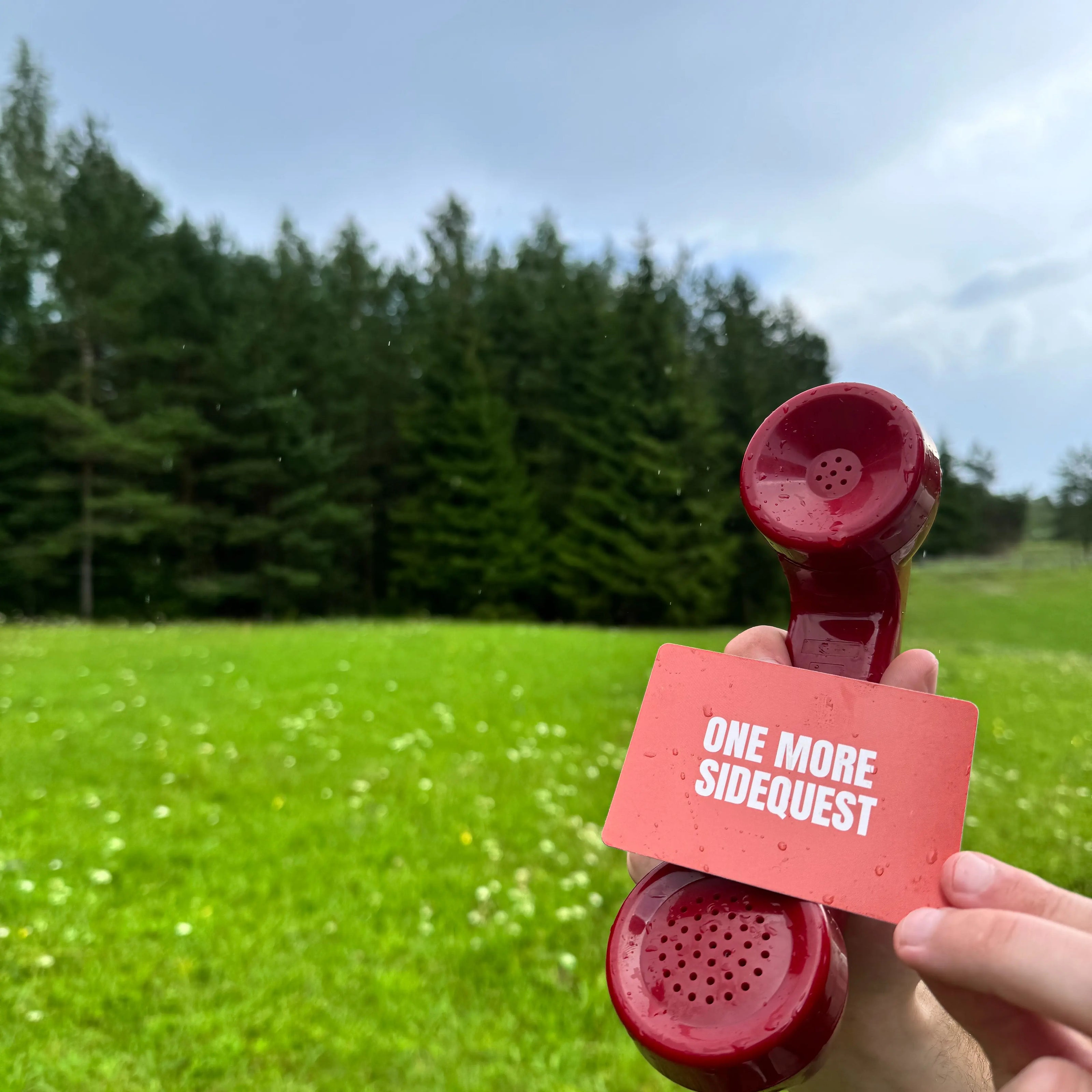 Red phone receiver with a card labeled 'One More Sidequest' held against a green field and trees.