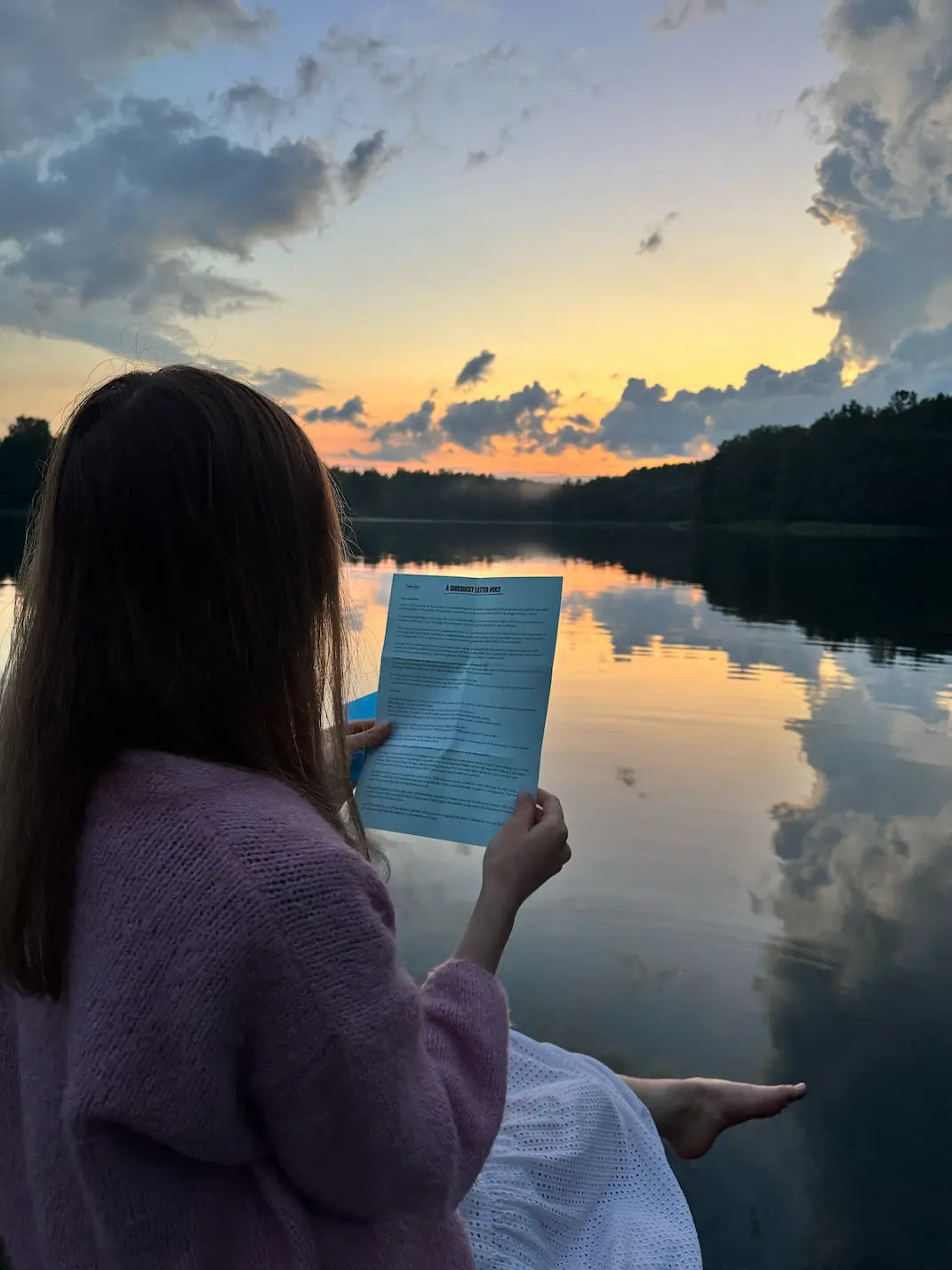 Person reading a letter by a lake at sunset