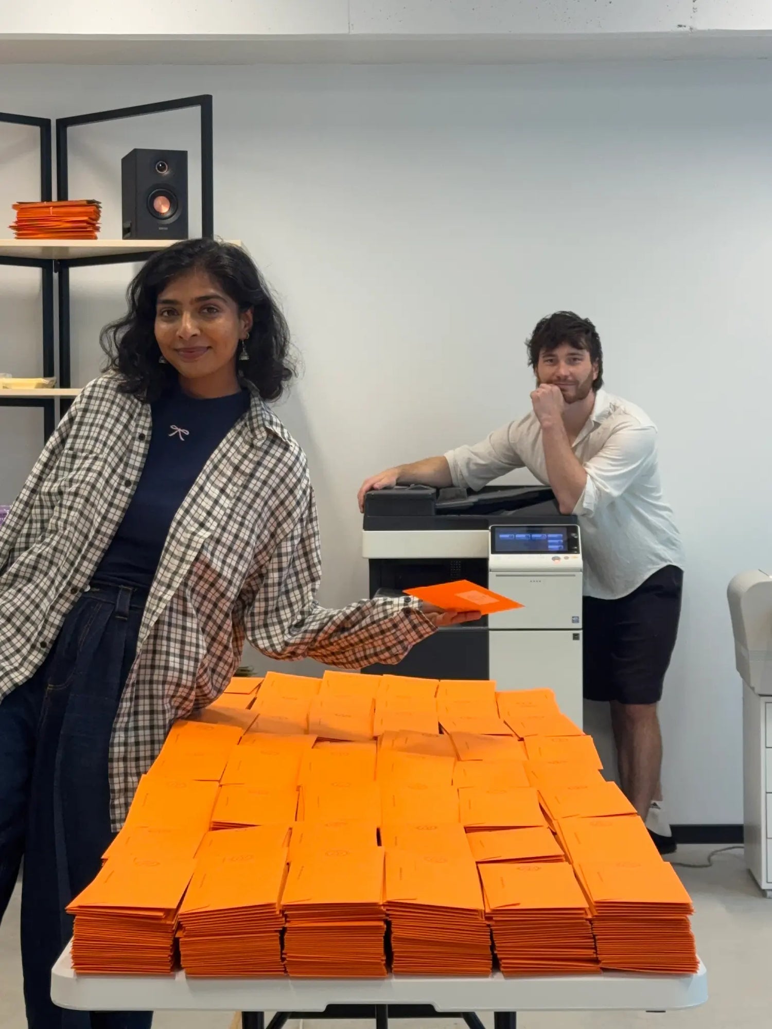 A woman in a plaid blazer holds an orange envelope next to a table stacked with orange envelopes, with a man working at a printer in the background.