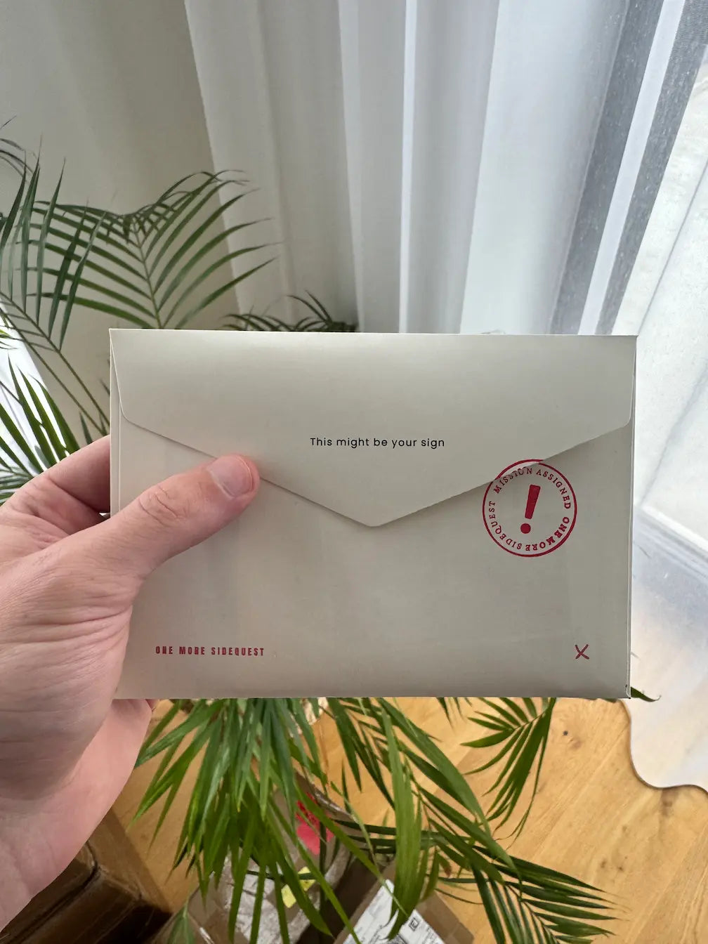 Hand holding a Sidequest Mail Club envelope with a red mission stamp before opening.